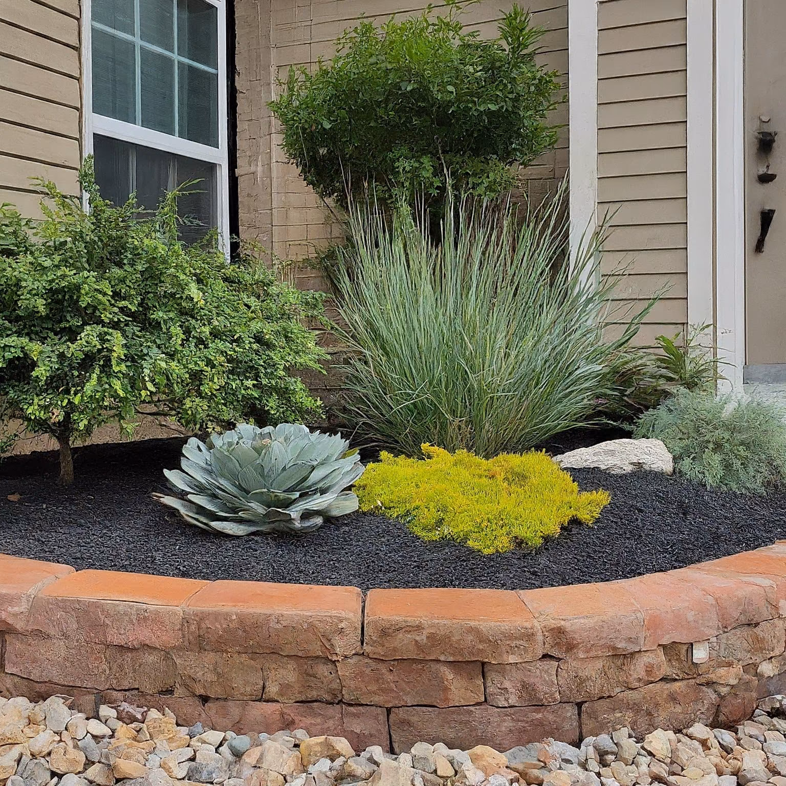 A raised stone planter bed with agave, ornamental grasses, and gravel groundcover