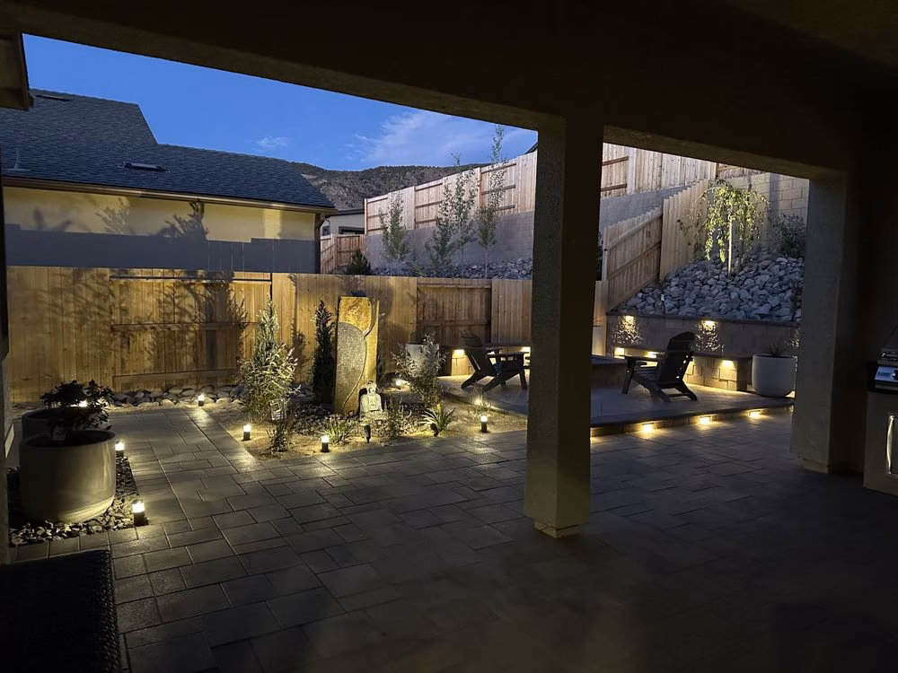 A covered patio at dusk with a lit fire feature, paver flooring, and string lighting