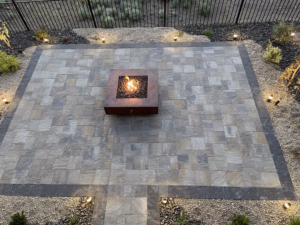 A paver patio with a built-in gas fire pit table, edged with landscape lighting and gravel