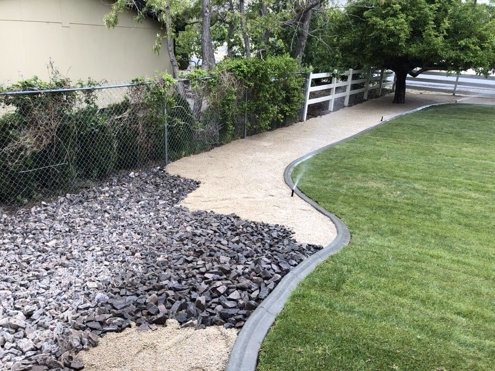 Crushed rock and pea gravel bordering a lawn along a chain-link and white ranch fence
