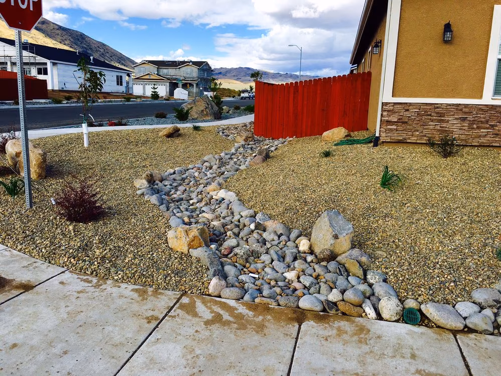 A dry creek bed of river rock winding through a gravel yard beside a red wood fence