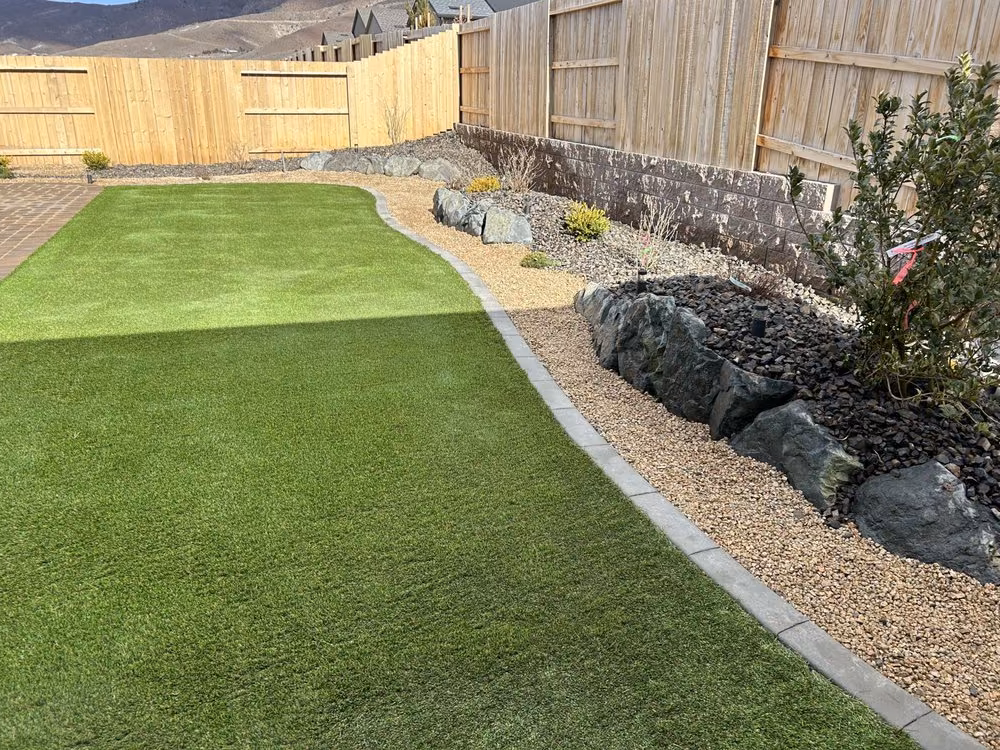 Artificial turf bordering natural lawn, divided by a paver edge, beside a rock and boulder bed